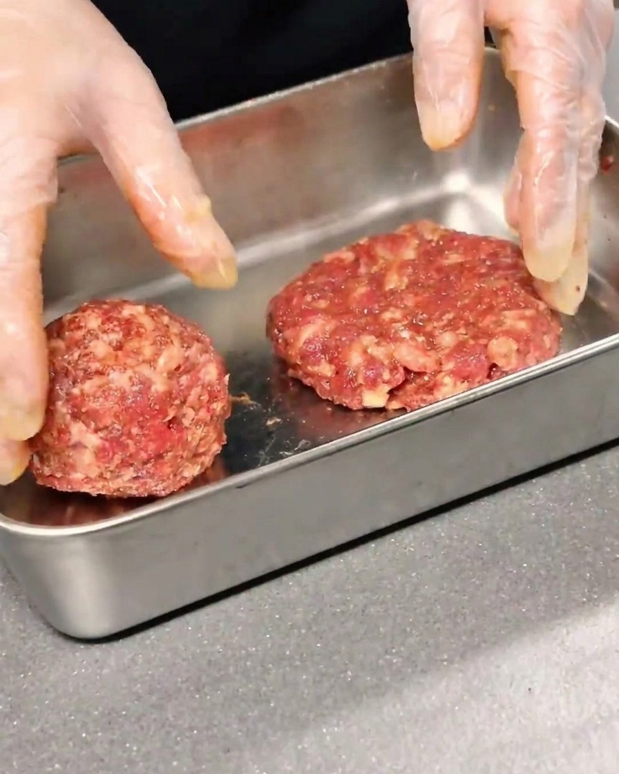 Dos discos de carne sazonada siendo moldeados y aplanados en un recipiente metálico.