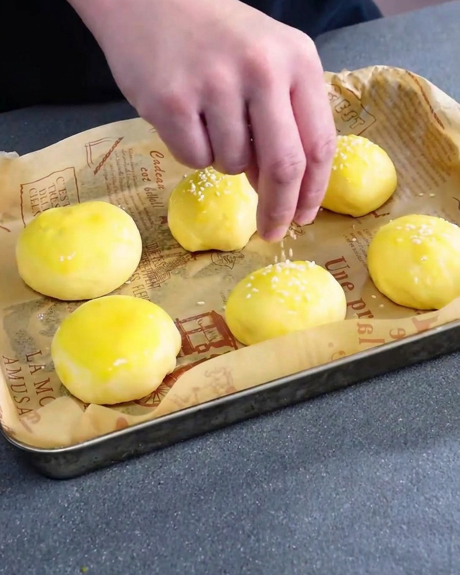 Semillas de sésamo blanco esparciéndose sobre los panes de hamburguesa crudos y pintados con huevo.
