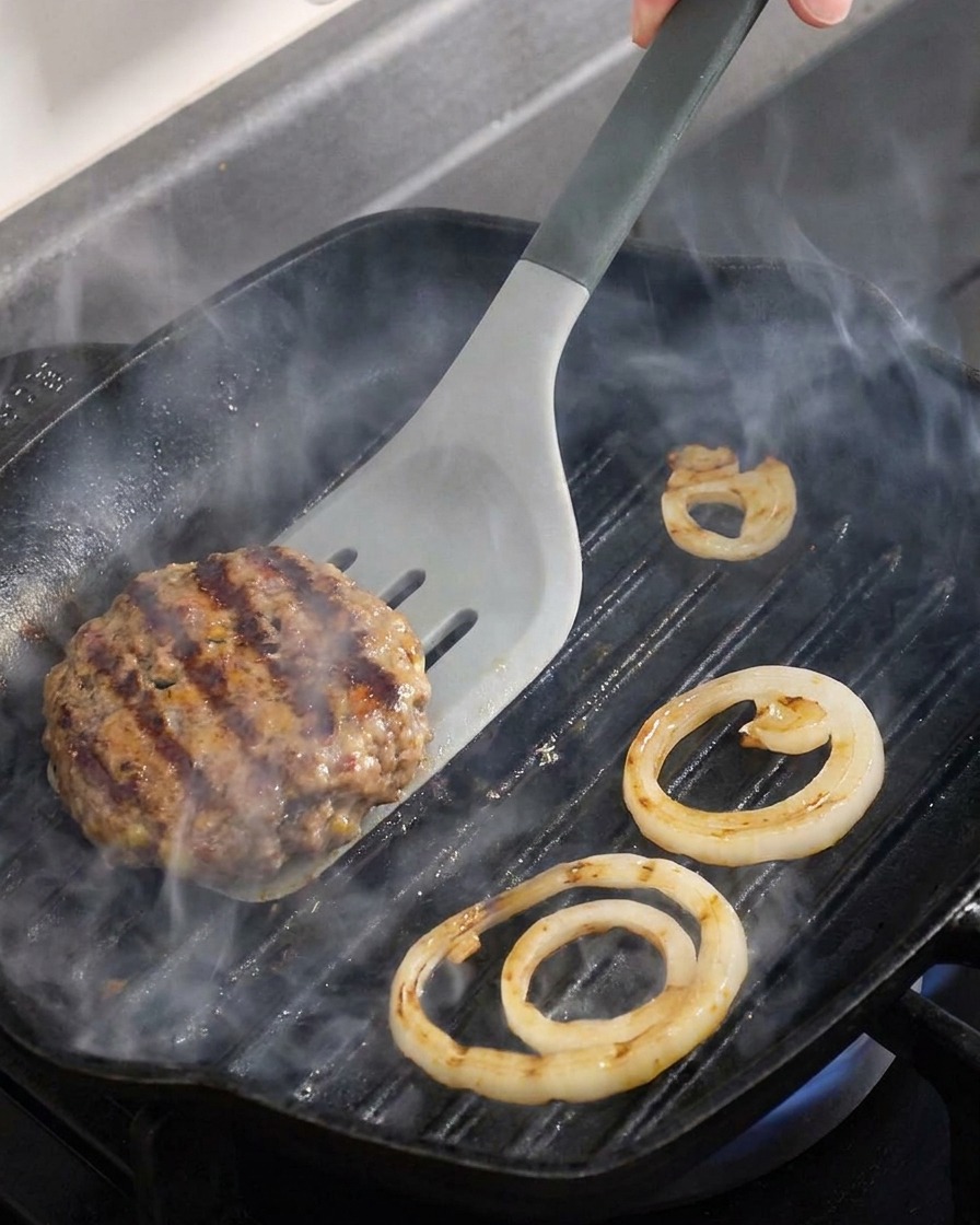 Primer plano de una hamburguesa de carne y aros de cebolla dándoles la vuelta en una plancha caliente.