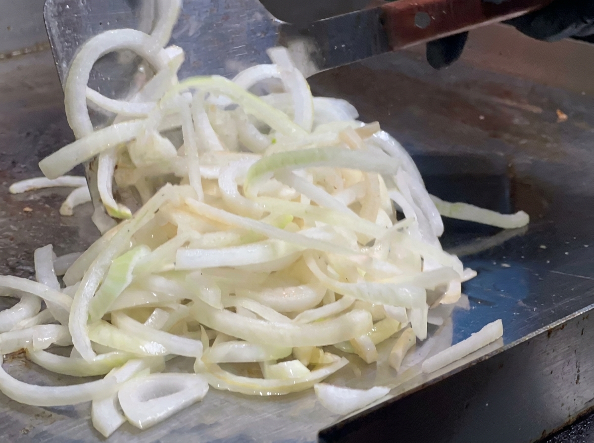 Tiras de cebolla siendo extendidas por una plancha caliente usando una espátula de metal grande.