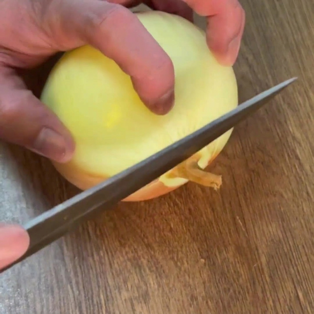 Manos cortando los extremos de una cebolla pelada sobre una tabla de madera.