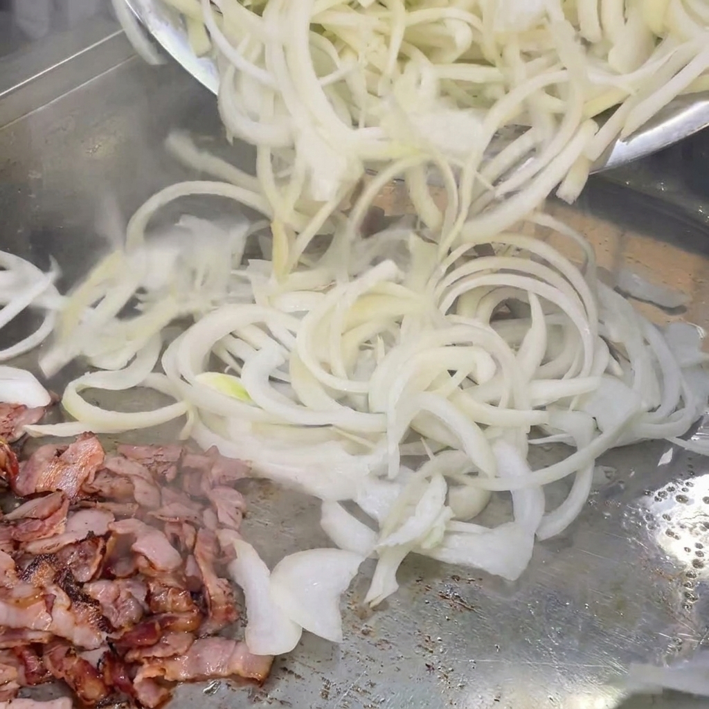 Cebollas blancas en láminas siendo mezcladas con los trozos de beicon en una plancha caliente.