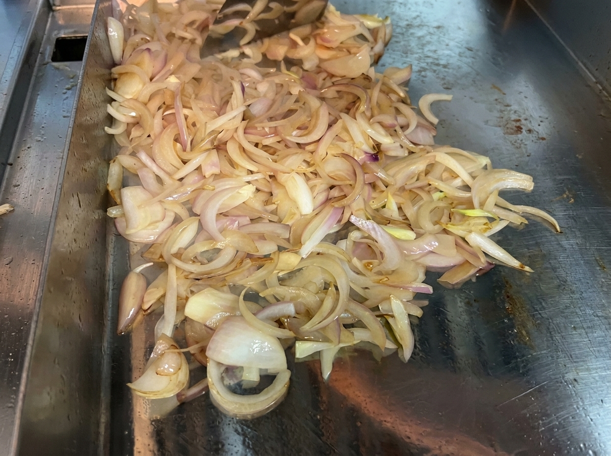 Cebollas en rodajas siendo removidas continuamente con una espátula sobre una superficie caliente para ablandar y caramelizar.