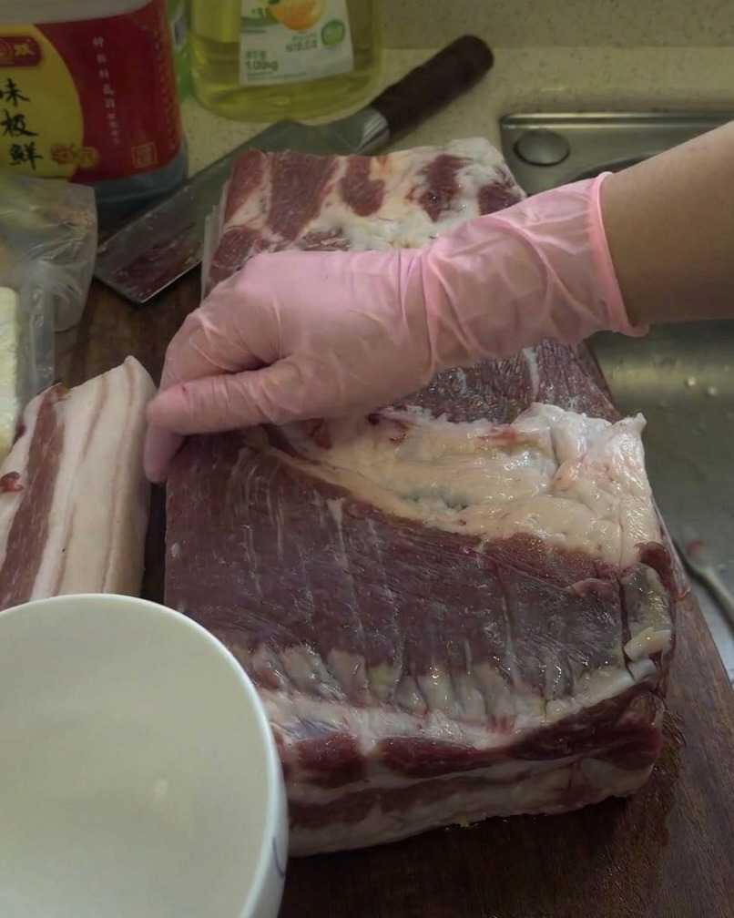 Un trozo grande de panceta cruda siendo frotado con licor blanco sobre una tabla de cortar de madera.
