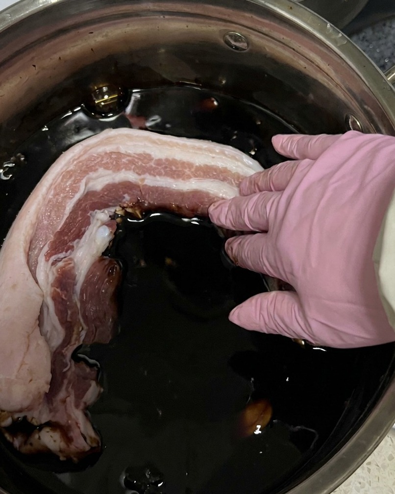 Tiras de panceta cruda siendo sumergidas en una olla con adobo de salsa de soja oscura.