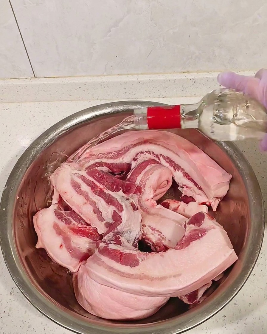 Trozos de panceta cruda en un bol de metal siendo rociados con licor blanco transparente.