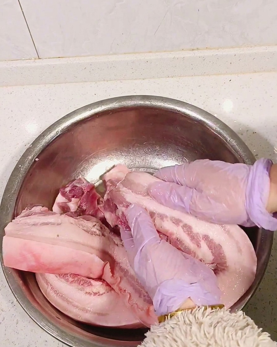 Manos con guantes morados masajeando licor blanco en la panceta cruda.