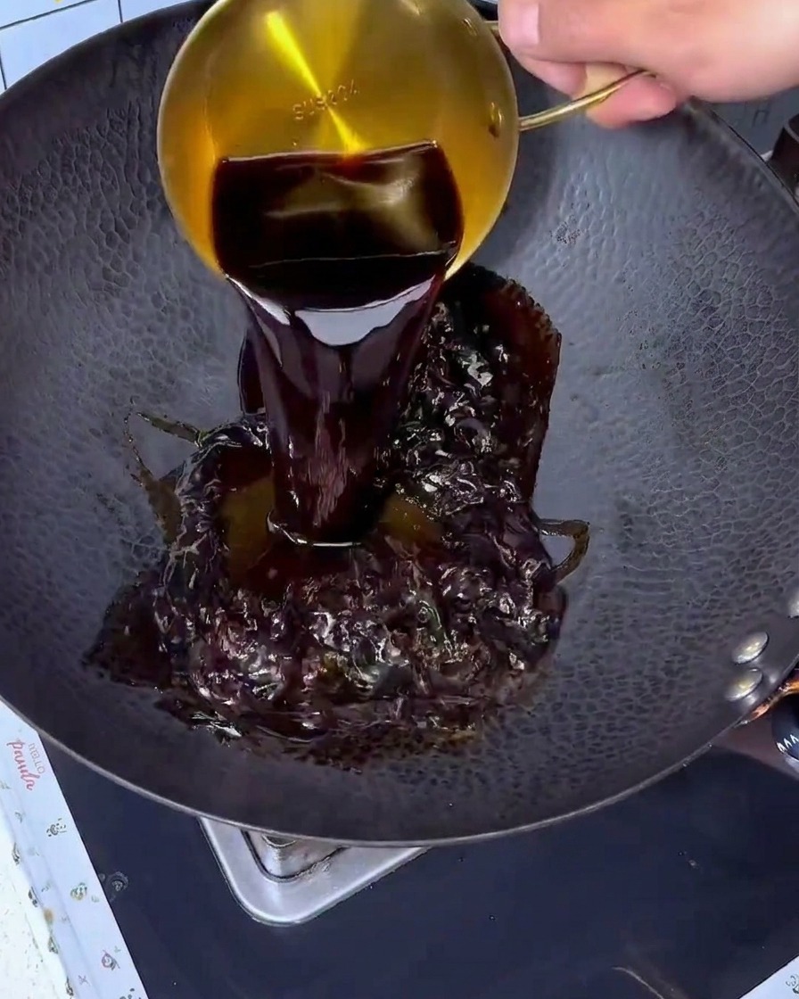 Salsa de soja siendo vertida desde una taza medidora dorada en un wok negro grande.