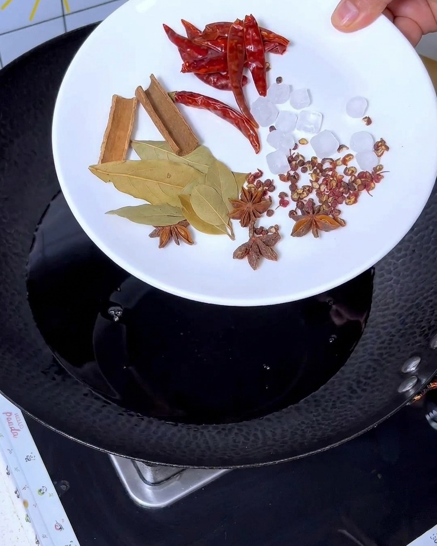 Especias como canela, anís estrellado, hojas de laurel, chile y azúcar cande siendo añadidas al líquido oscuro en el wok.