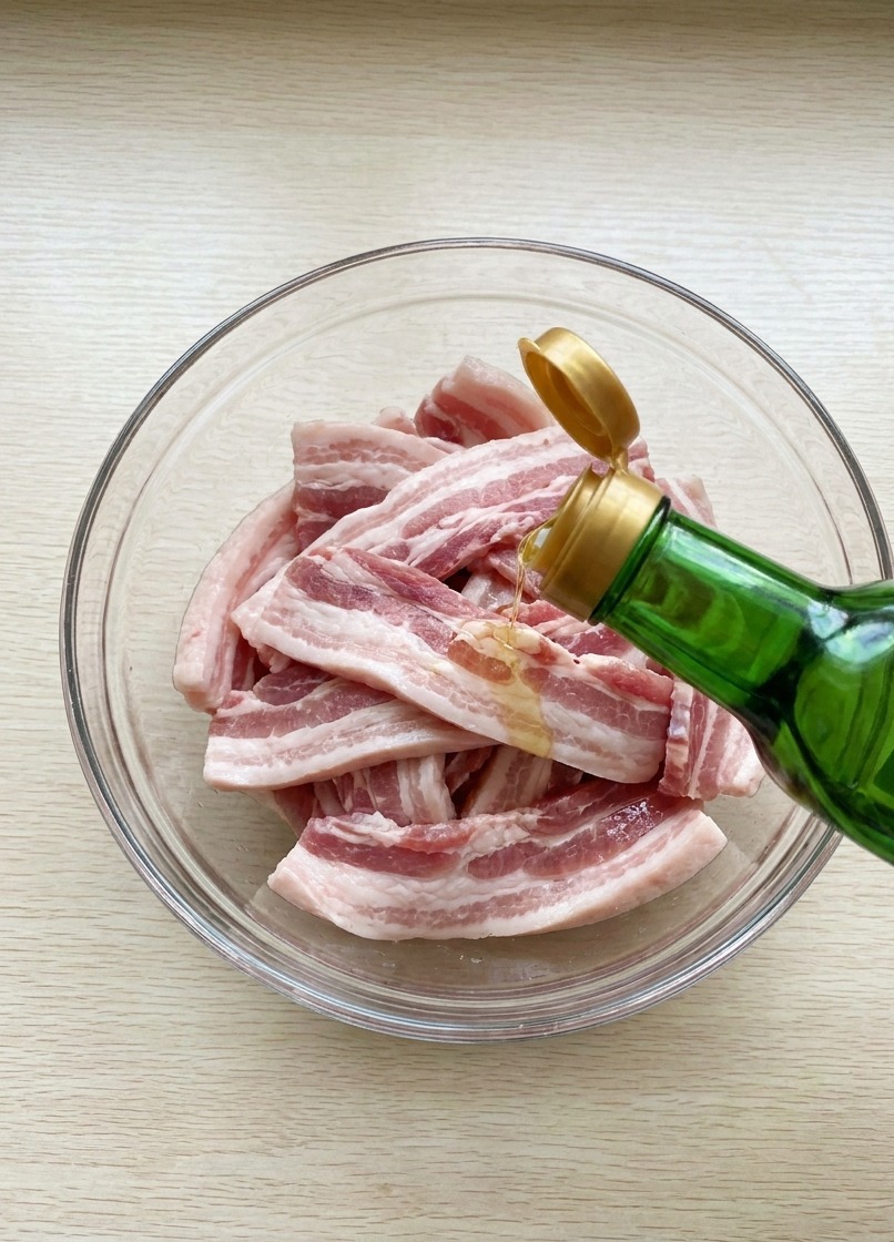 Salsa de soja vertiéndose desde una botella de cristal sobre las tiras de cerdo crudas en un cuenco.