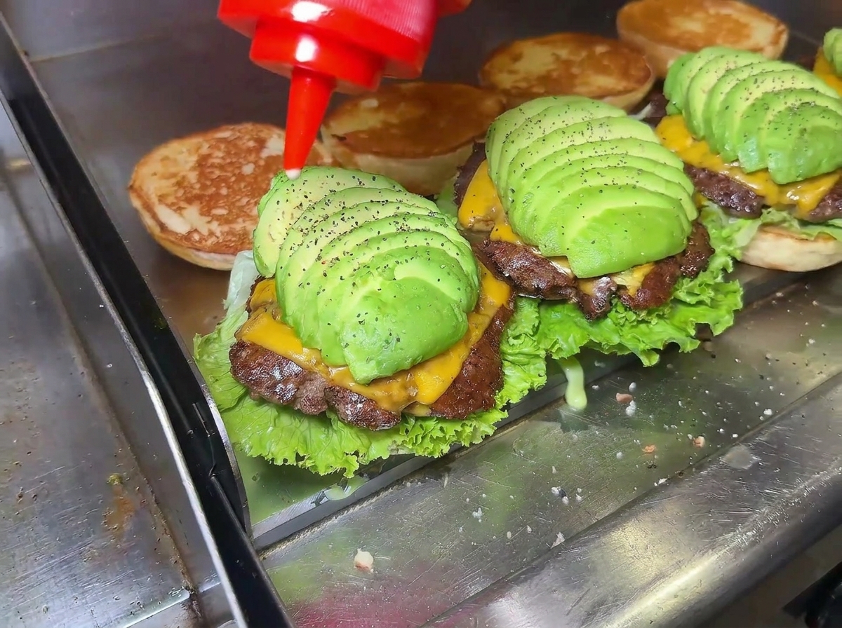Primer plano de una mano echando salsa sobre una hamburguesa de aguacate perfectamente montada.