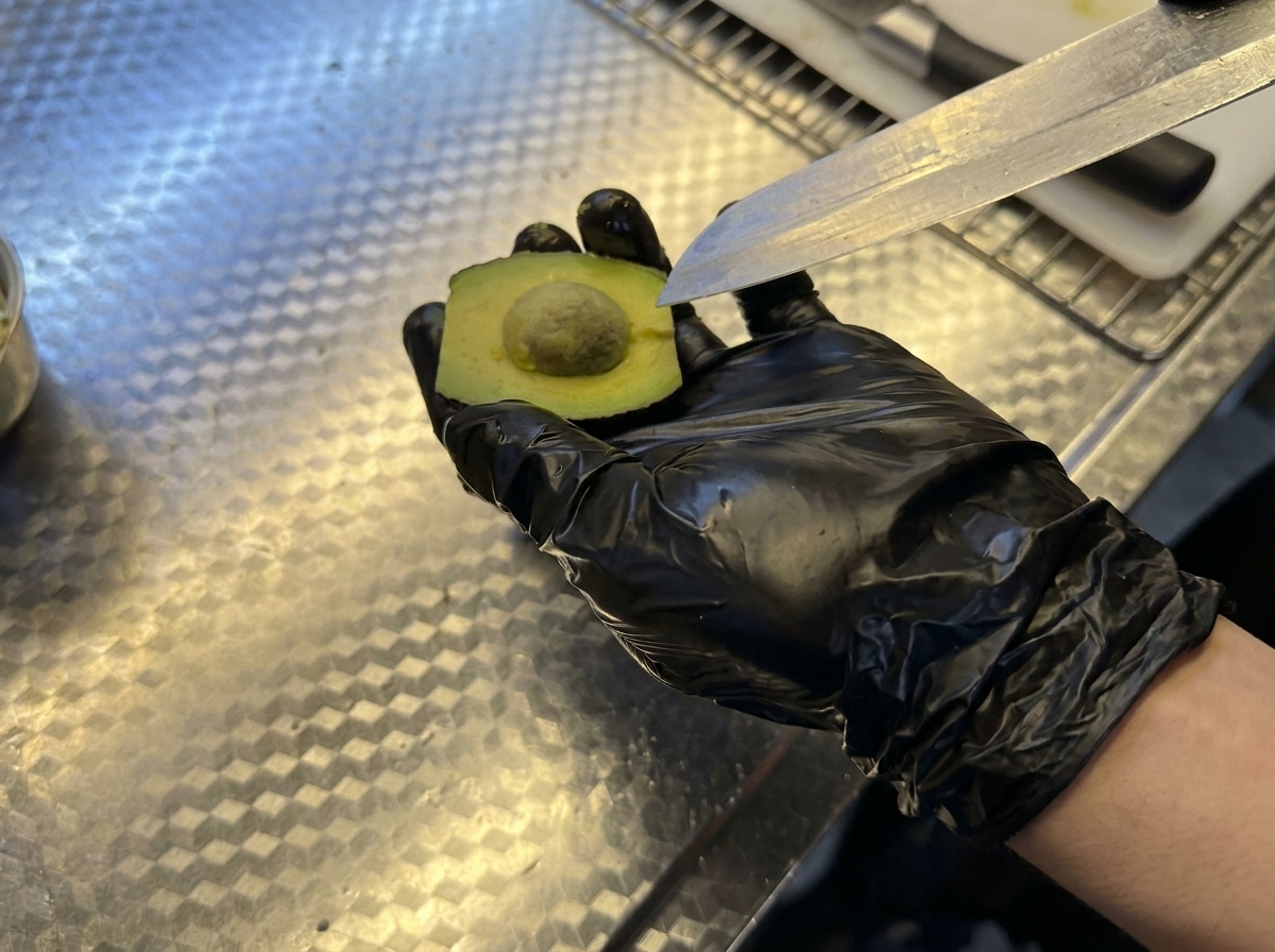 Mano con guante negro sosteniendo medio aguacate mientras se retira el hueso con un cuchillo.