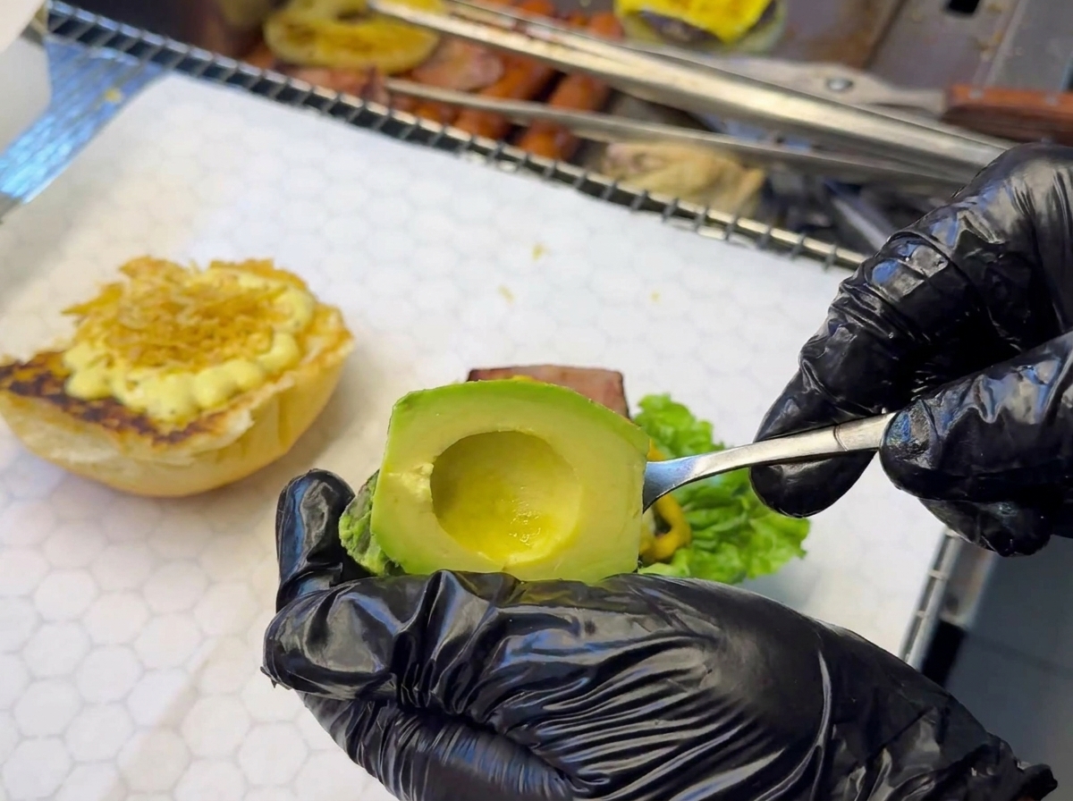 Mano con guante negro colocando láminas de aguacate sobre la base de la hamburguesa.