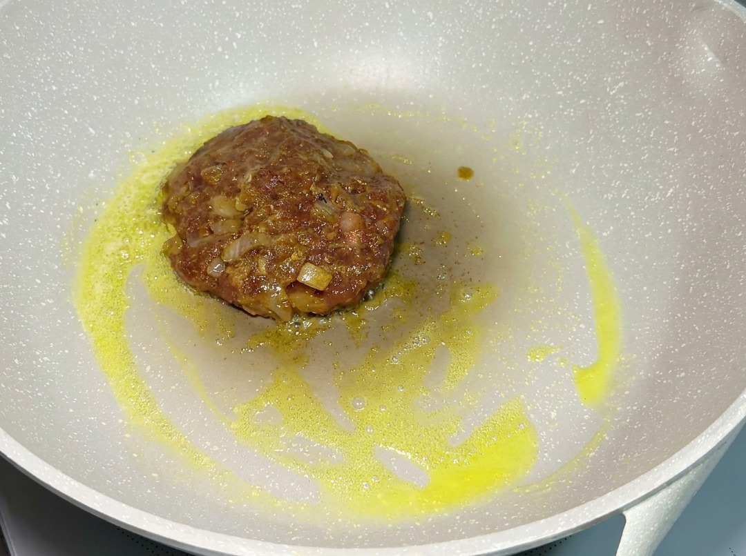 Una hamburguesa de ternera cruda sazonada friéndose en una sartén clara a fuego lento, rodeada de grasa derretida.