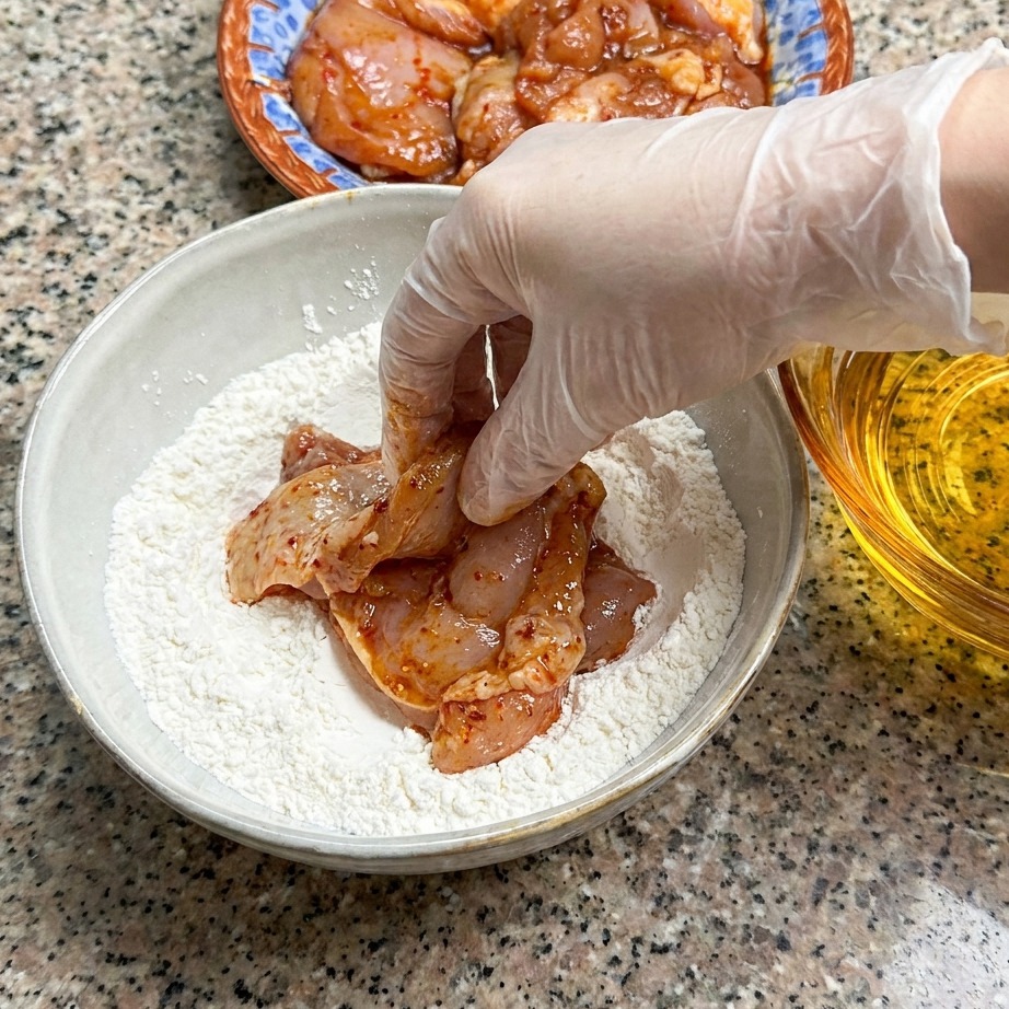 Un muslo de pollo marinado crudo siendo presionado en un cuenco con mezcla de harina seca.