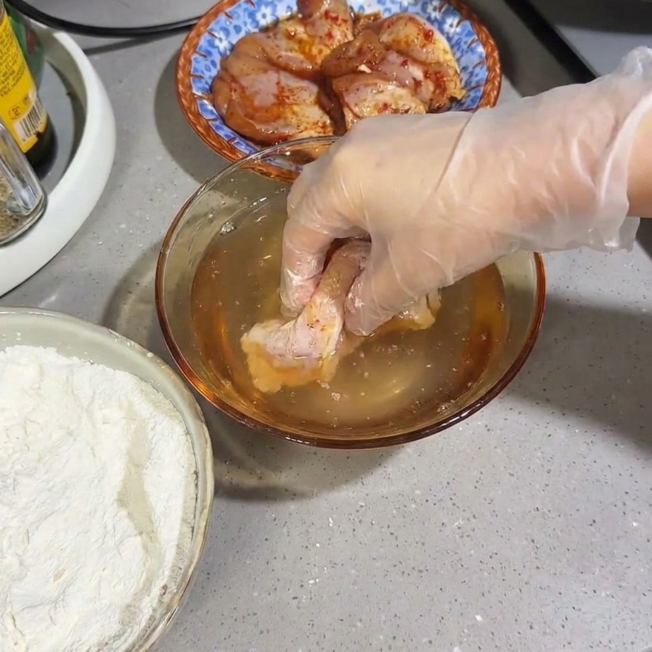 Un muslo de pollo crudo cubierto de harina siendo sumergido brevemente en un cuenco de agua.