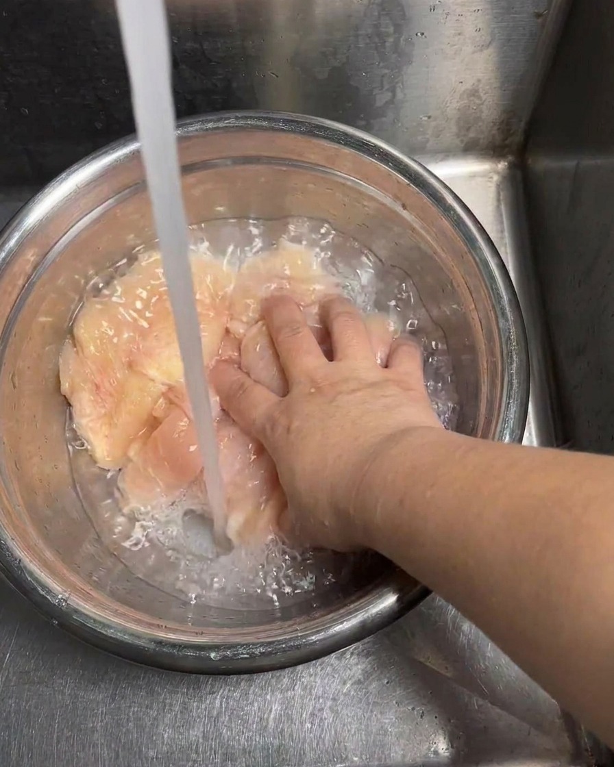 Láminas de pollo crudo siendo enjuagadas bajo un chorro constante de agua fría.