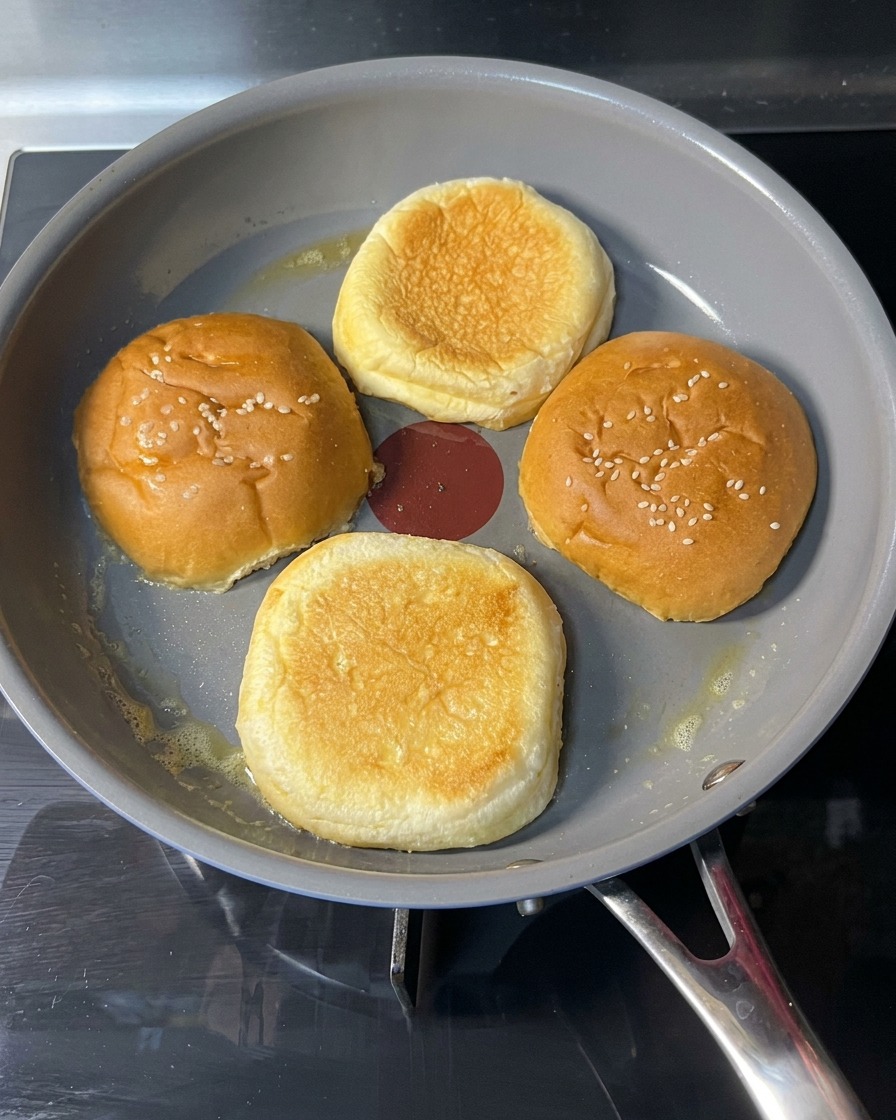 Una mano volteando un pan de hamburguesa tostado en la sartén, mostrando una superficie dorada y crujiente.