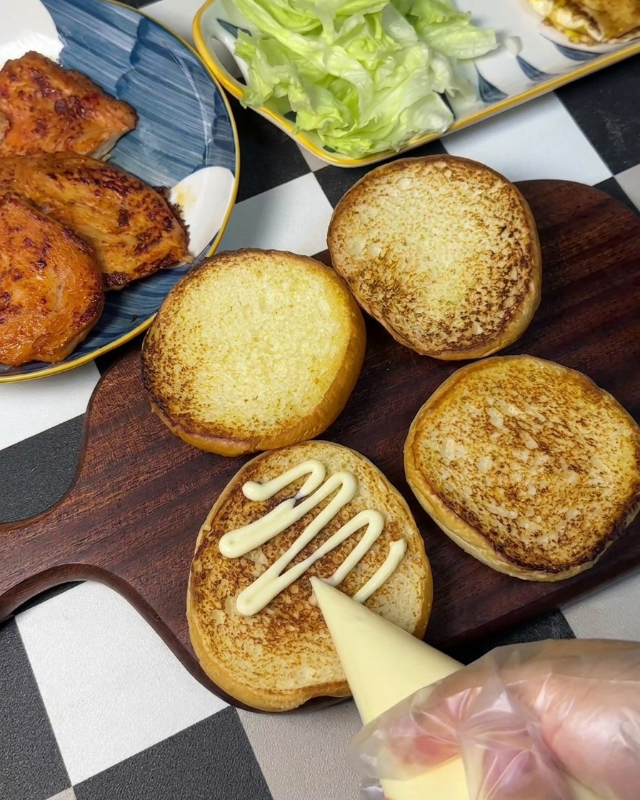 Una mano usando una manga pastelera para poner mayonesa en un patrón en zigzag sobre un pan de hamburguesa tostado.