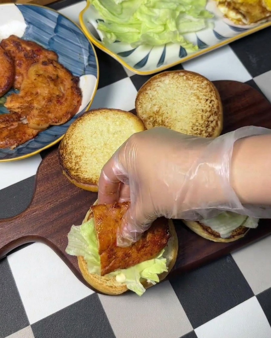 Una mano con guante colocando una pieza de pollo frito sobre una cama de lechuga fresca en el pan.