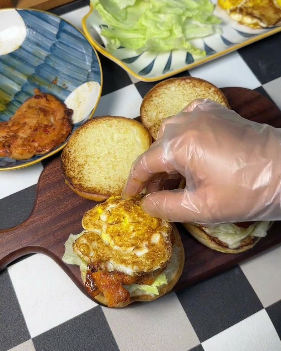 Una mano con guante colocando un huevo frito con bordes crujientes sobre la pieza de pollo.