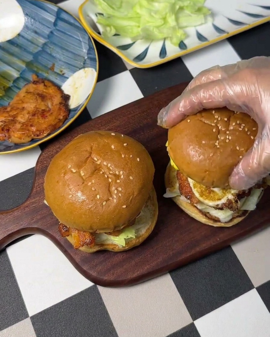 Una mano con guante colocando la tapa del pan sobre la pila de huevo, pollo y lechuga para completar el sándwich.