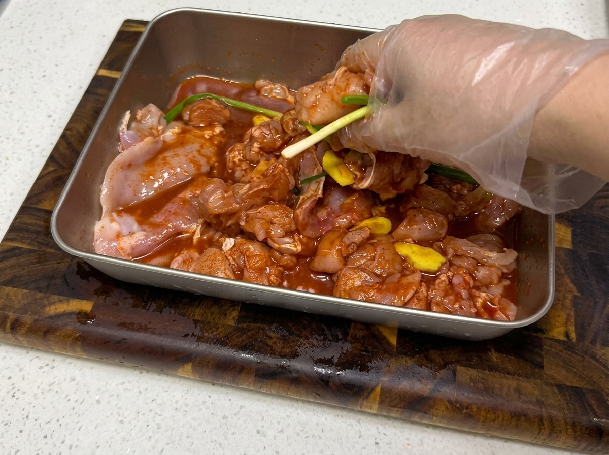 Manos mezclando el marinado con el pollo troceado en una bandeja metálica.