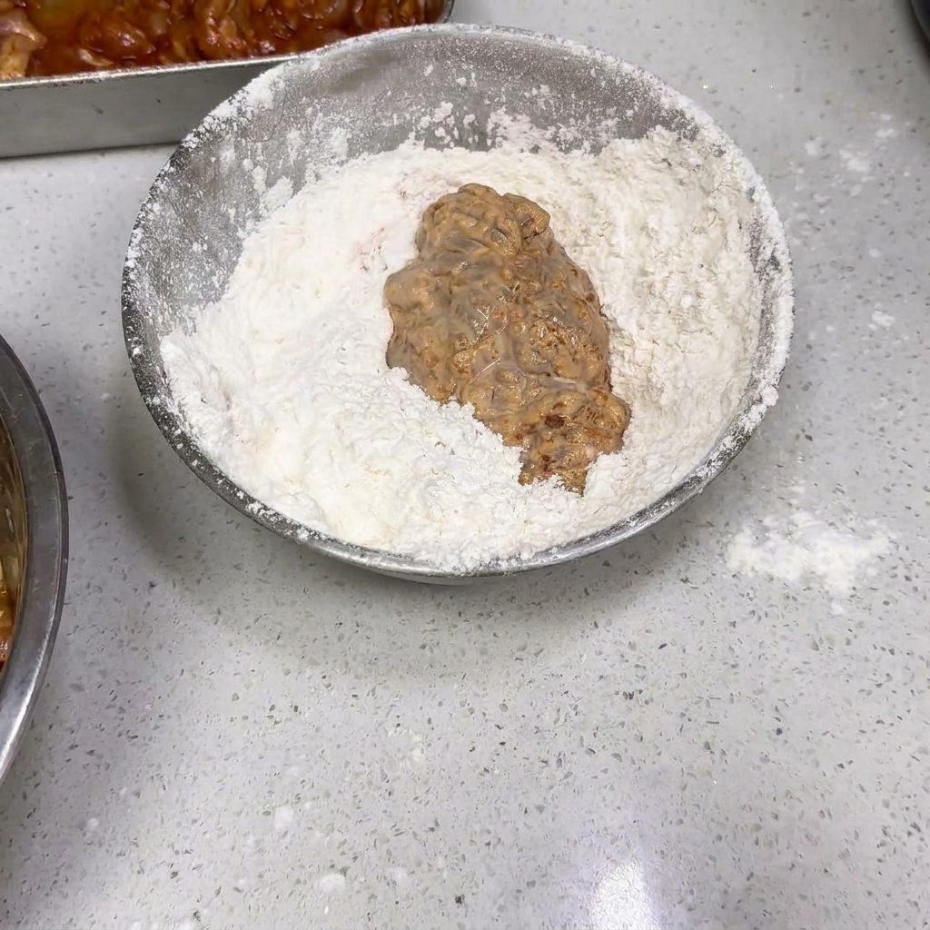 Un trozo de pollo humedecido siendo rebozado en un cuenco con harina seca.