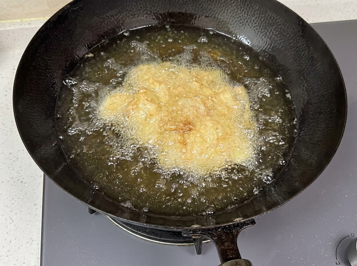 Trozos de pollo rebozados burbujeando en aceite caliente dentro de un wok oscuro.