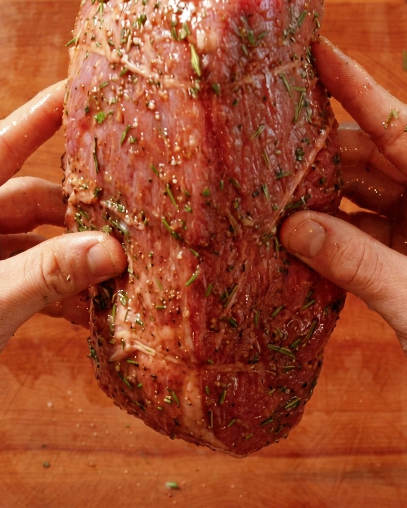 Manos colocando un roast beef crudo sazonado en una sartén caliente.