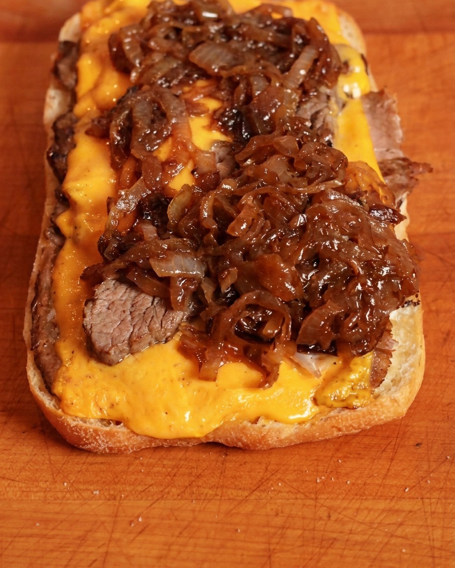 Cebollas caramelizadas de color oscuro puestas sobre el queso cheddar en la base del sándwich.