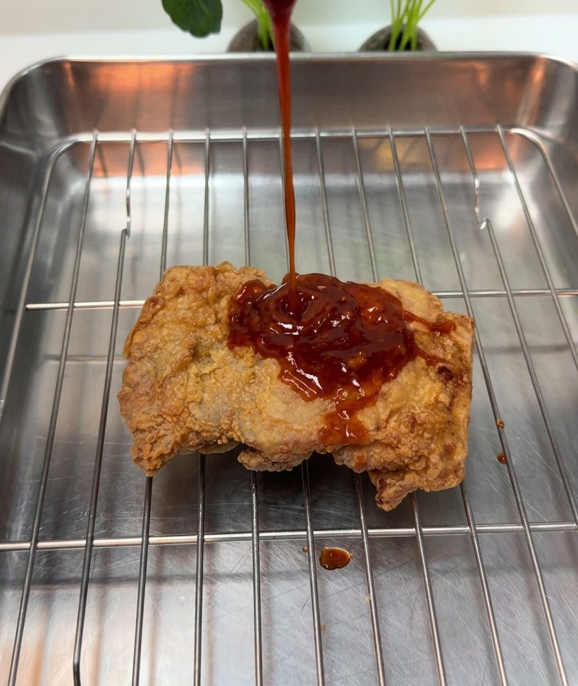 Salsa roja espesa siendo vertida sobre una pieza de pollo frito crujiente en una rejilla metálica.