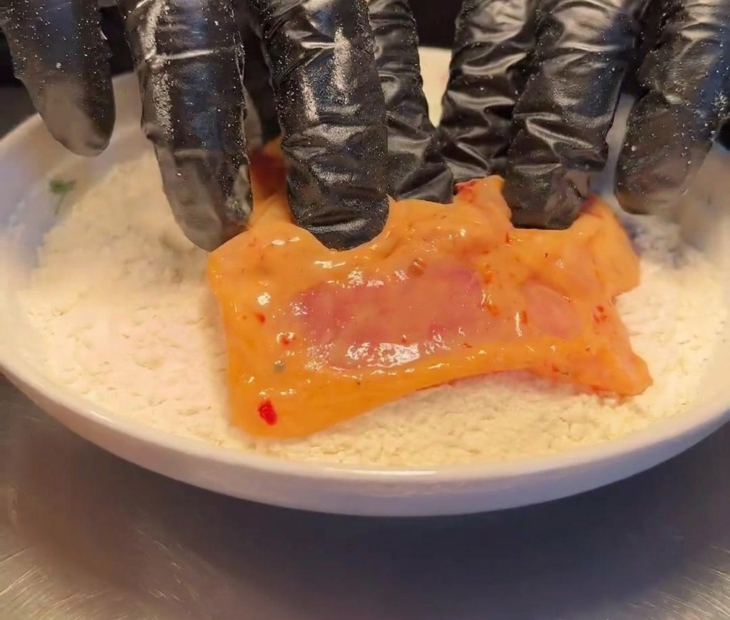 Un muslo de pollo crudo marinado siendo rebozado en un plato con harina blanca seca.