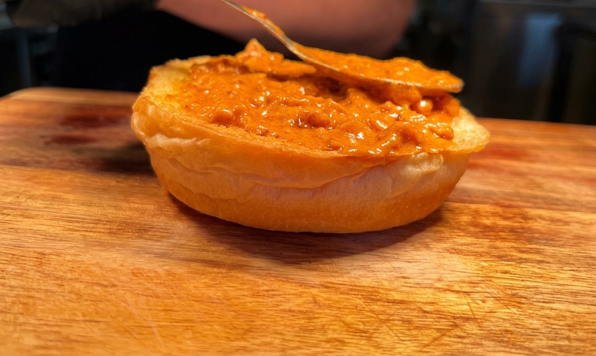 Una cuchara esparciendo salsa de crema picante naranja en la base tostada de un pan de hamburguesa.