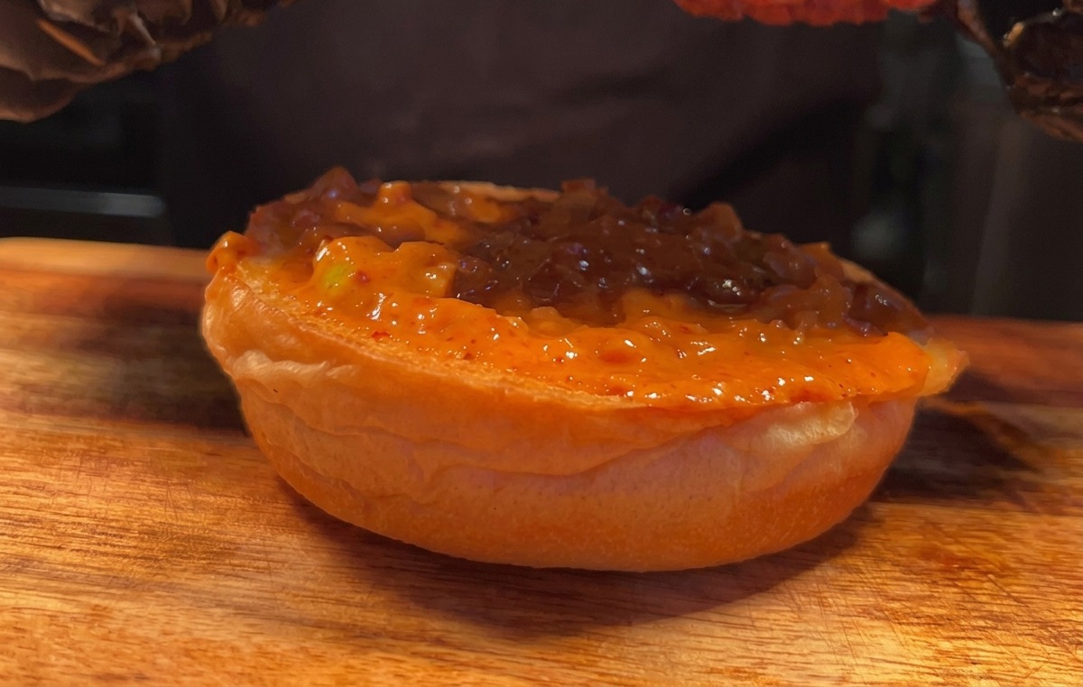 Una mano con guantes colocando un trozo de pollo frito crujiente sobre un pan cubierto de salsa.