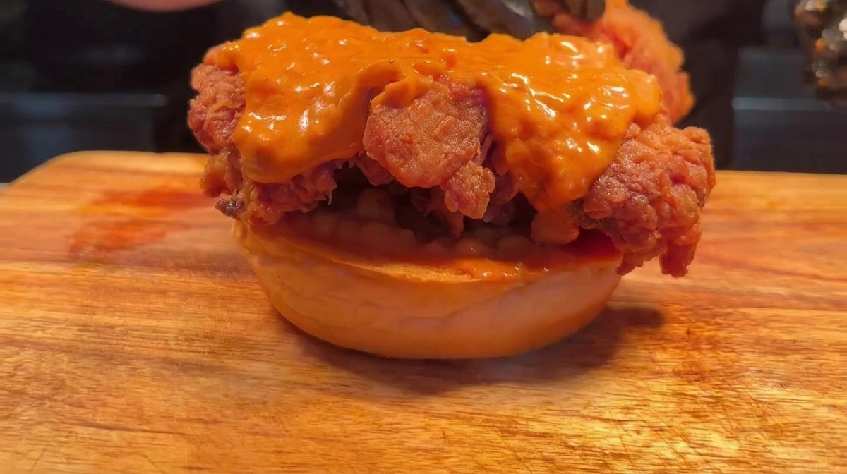 Manos con guantes armando una hamburguesa doble de pollo apilando un segundo trozo frito encima del primero.