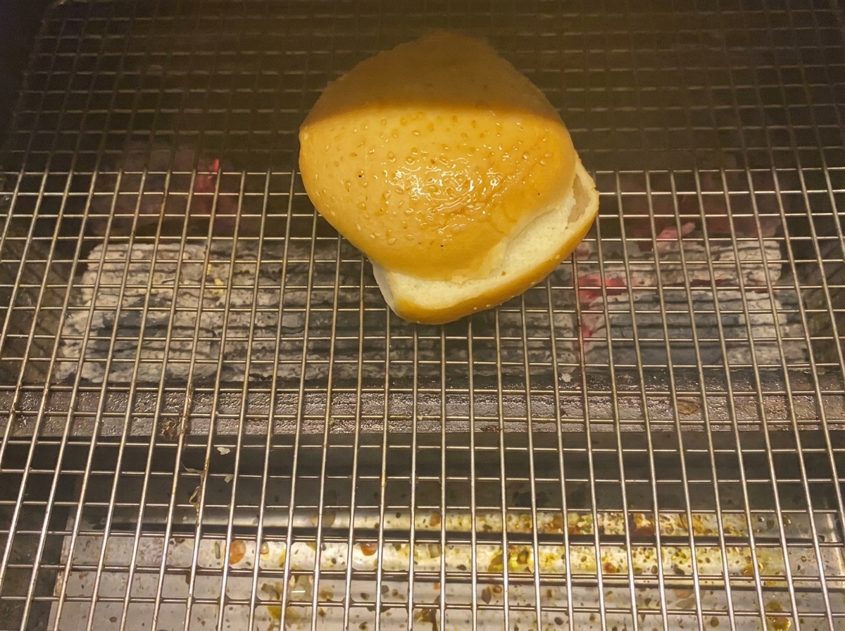 Una mitad de pan de hamburguesa tostándose boca abajo en una parrilla de carbón.