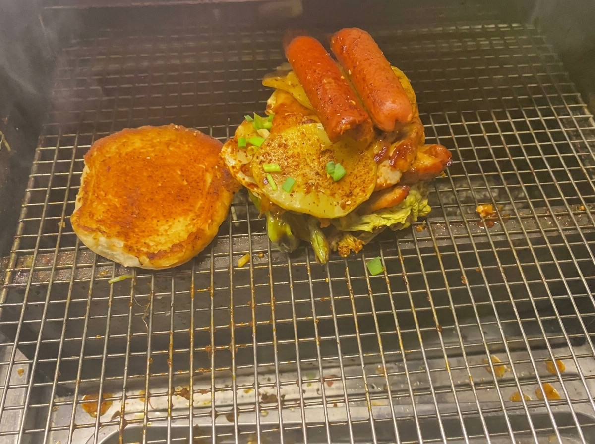 Una hamburguesa imponente cargada con carnes, patatas asadas y salchichas junto a un pan tostado en la rejilla de la parrilla.
