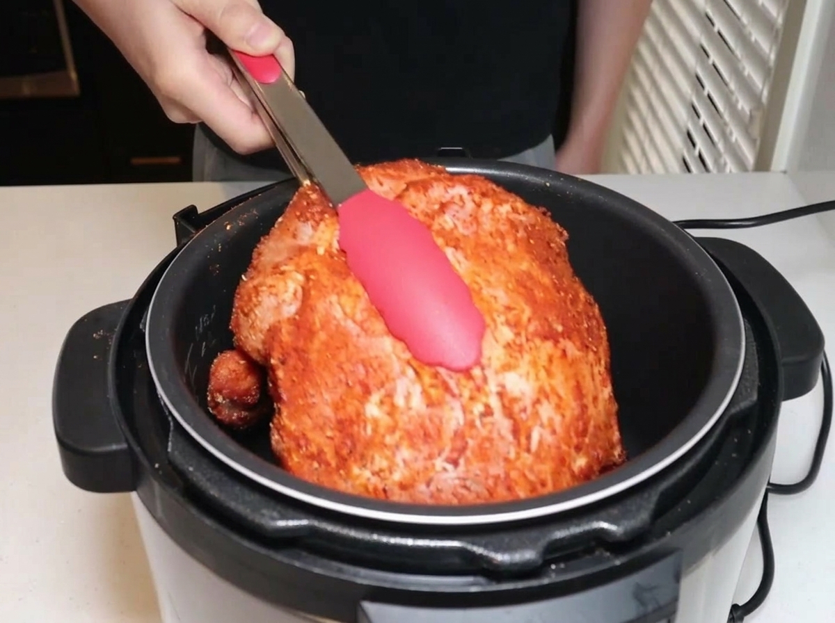 Paleta de cerdo sazonada siendo introducida en una olla a presión eléctrica con pinzas.