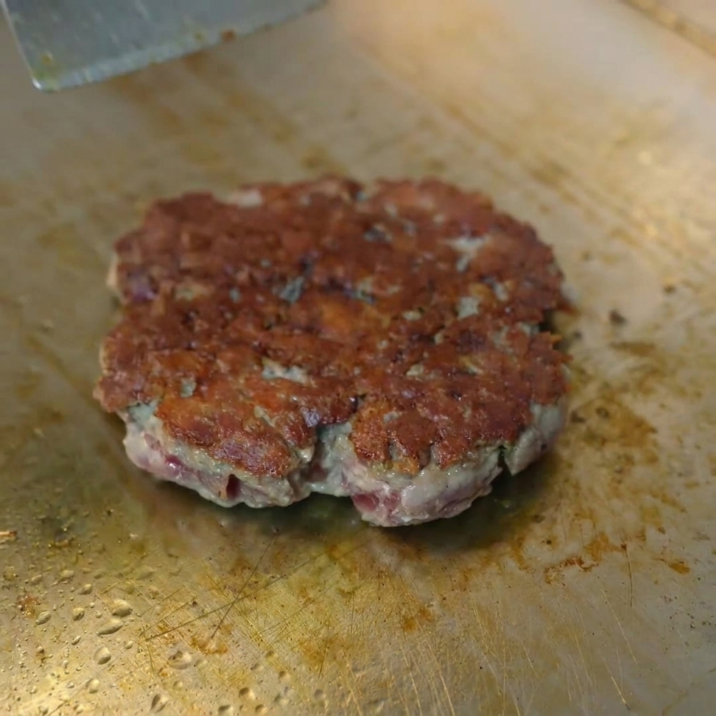 Una rebanada de queso amarillo fundiéndose sobre una hamburguesa dorada bajo una cúpula metálica.