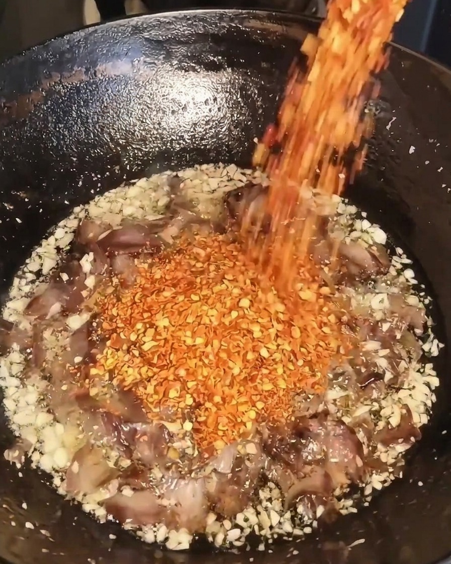 Hojuelas de chile rojo seco vertiéndose desde un recipiente a un wok que contiene ajo frito, carne y aceite.
