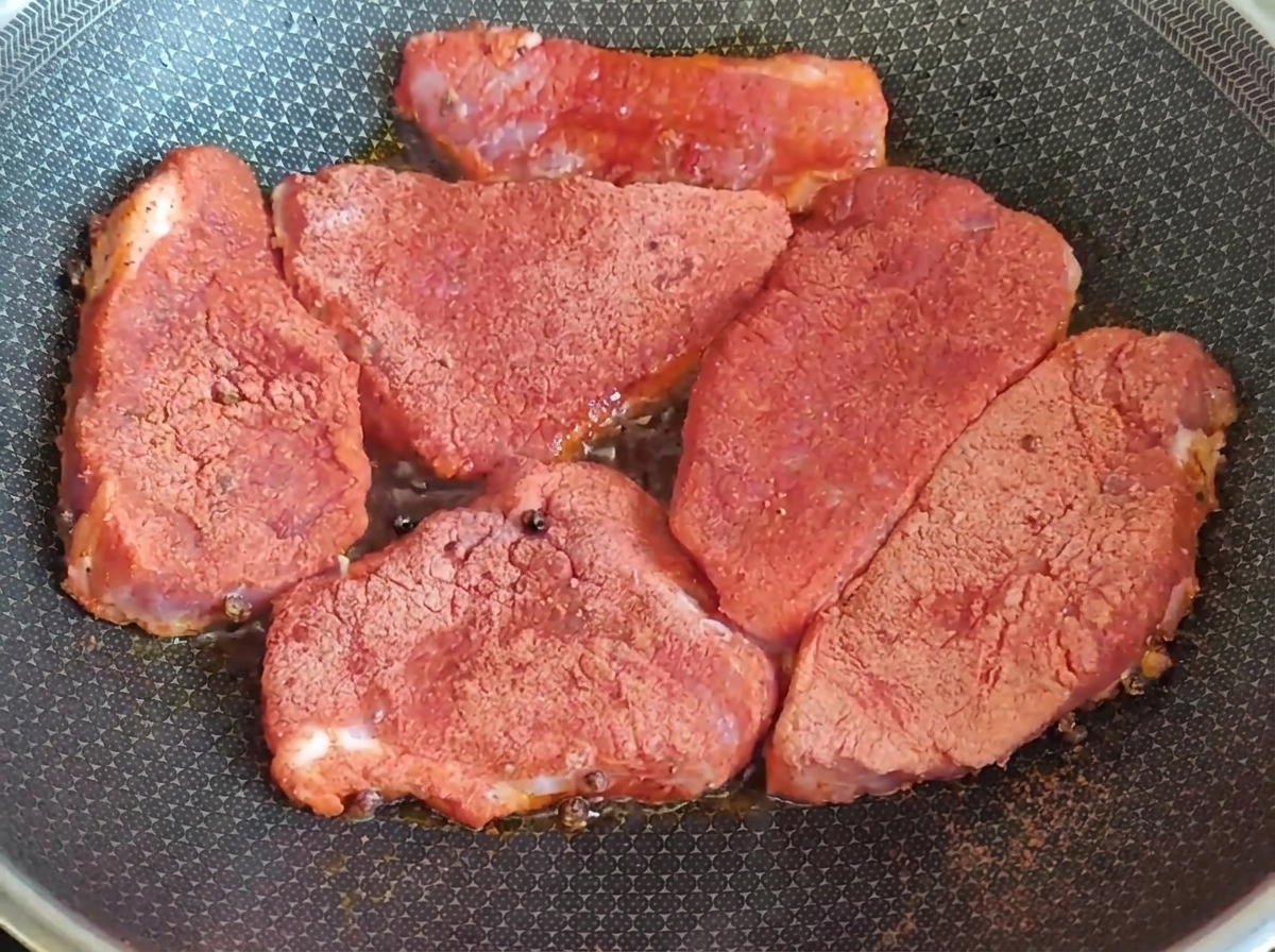 Trozos gruesos de carne cruda cubiertos con condimento rojo sellándose en una sartén caliente.