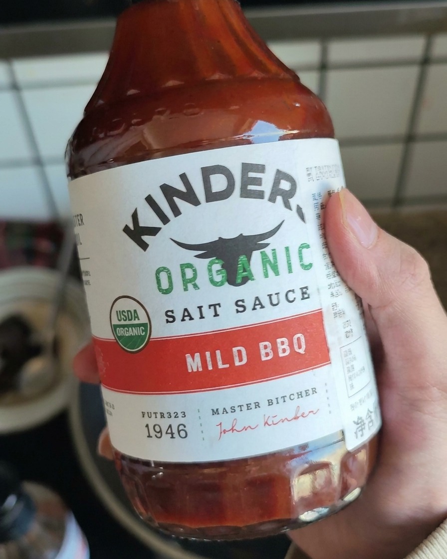 Una mano sosteniendo una botella de salsa barbacoa suave, preparándose para añadirla al cerdo desmenuzado.
