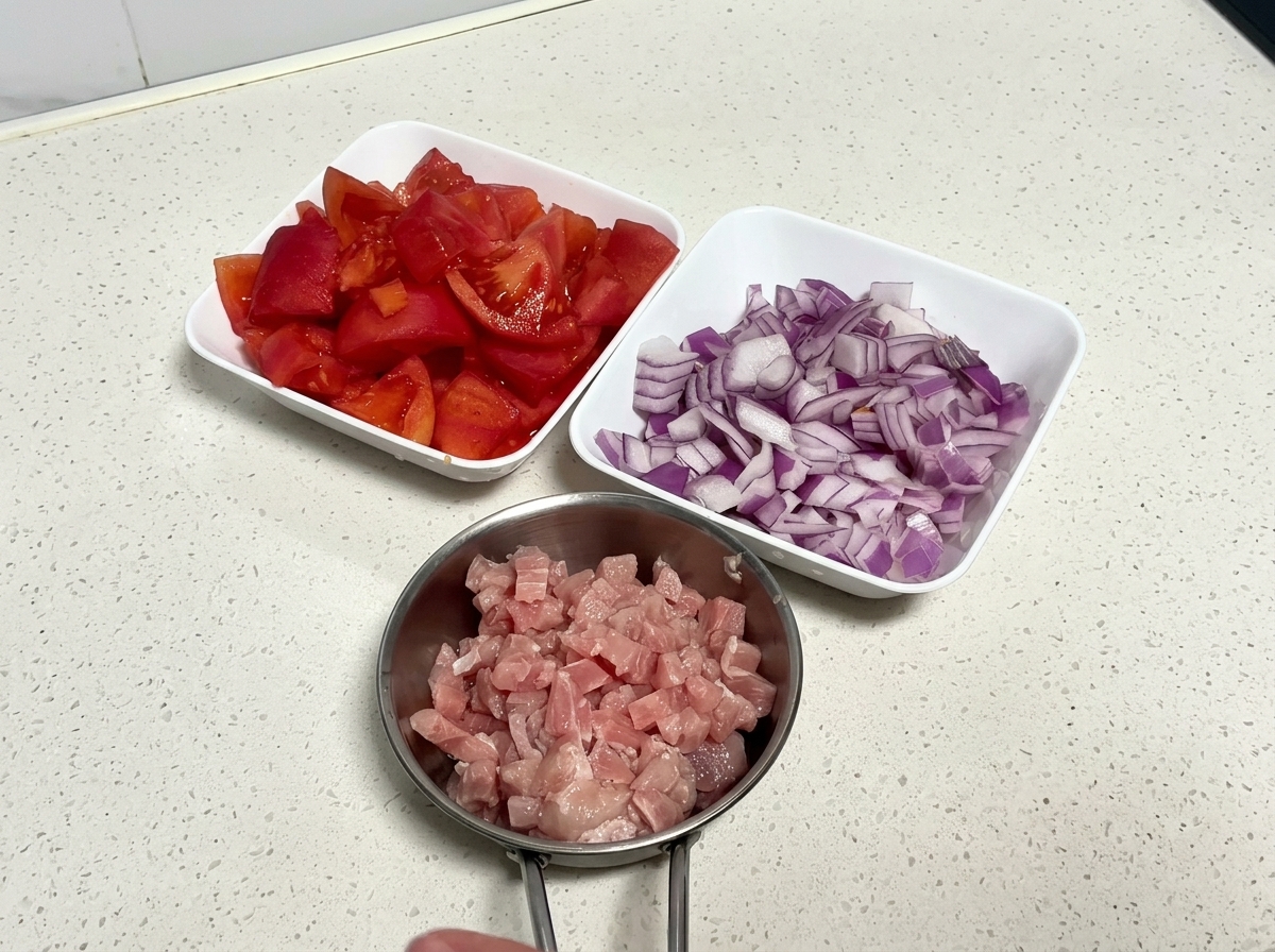 Vista cenital de tres cuencos blancos llenos de tomates troceados crudos, cebolla roja picada y cerdo en cubos crudos sobre una encimera limpia.