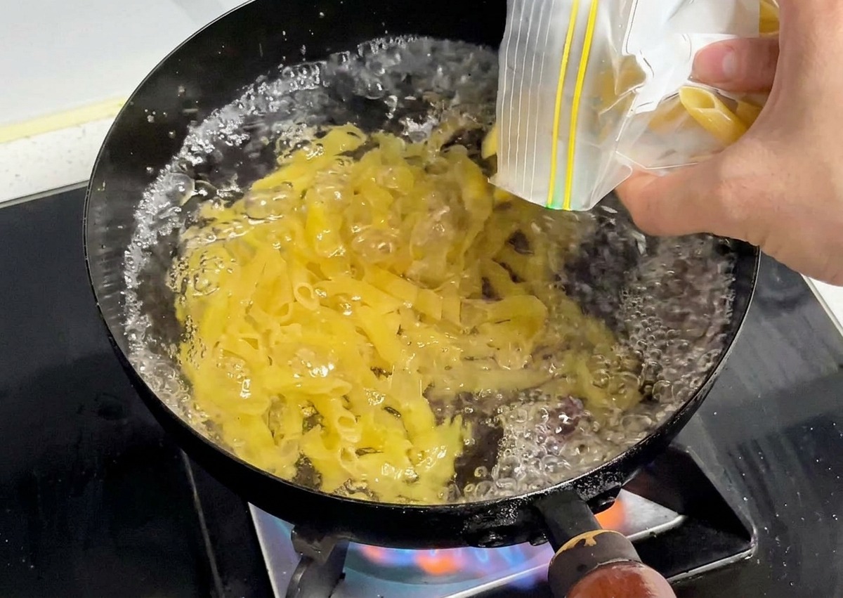 Pasta penne seca de color amarillo siendo vertida en una olla de agua hirviendo vigorosamente.