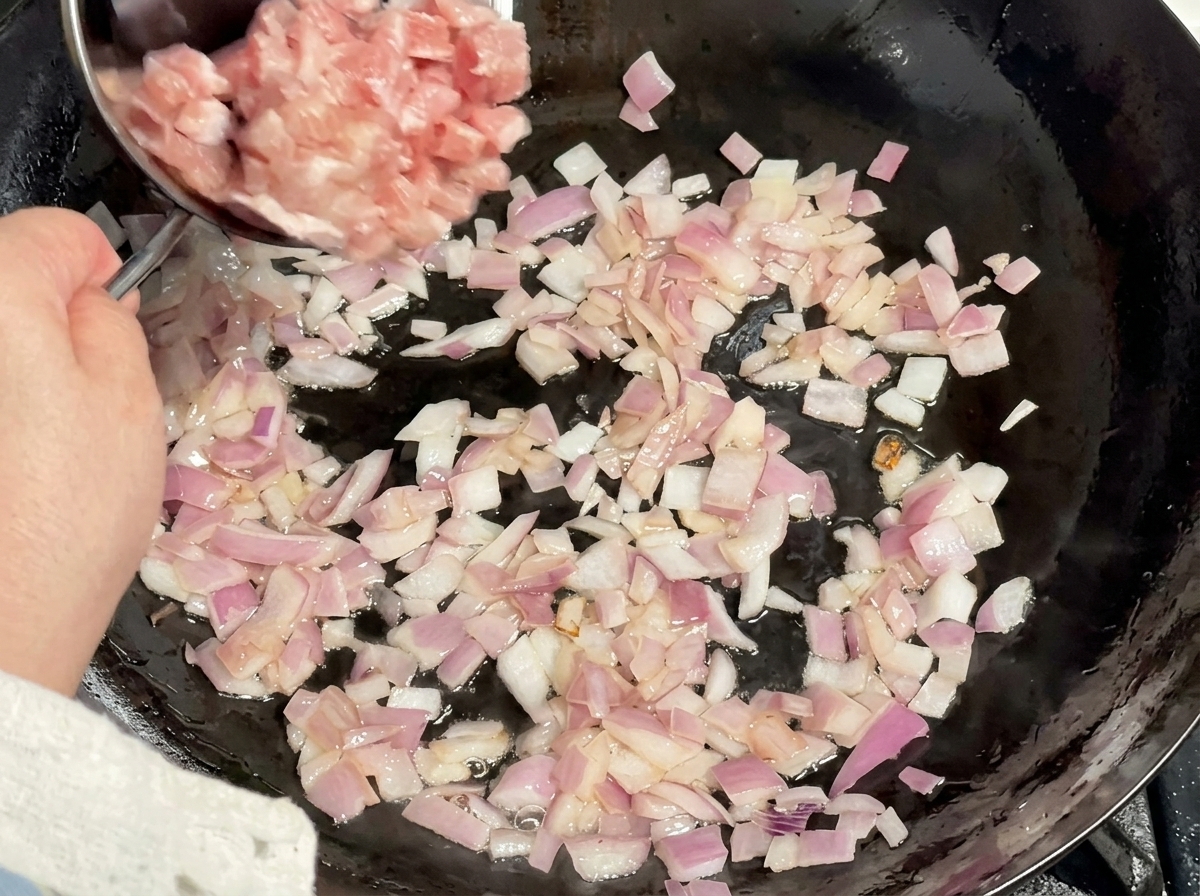 Cerdo en cubos crudo siendo vertido desde un cuenco de metal en un wok lleno de cebolla roja salteada.