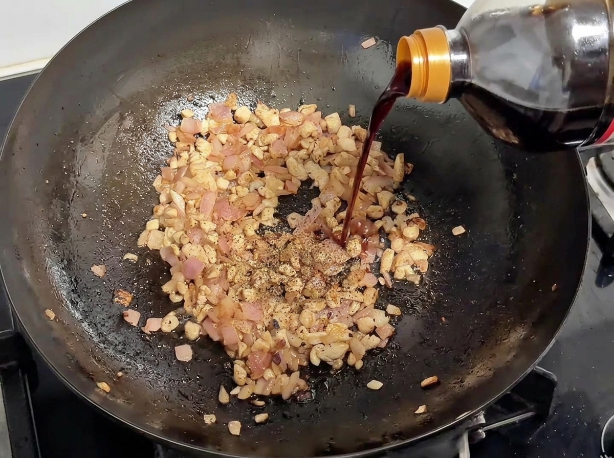 Salsa de soja siendo vertida desde una botella en un wok que contiene cerdo dorado y cebolla, cubierto con pimienta negra.