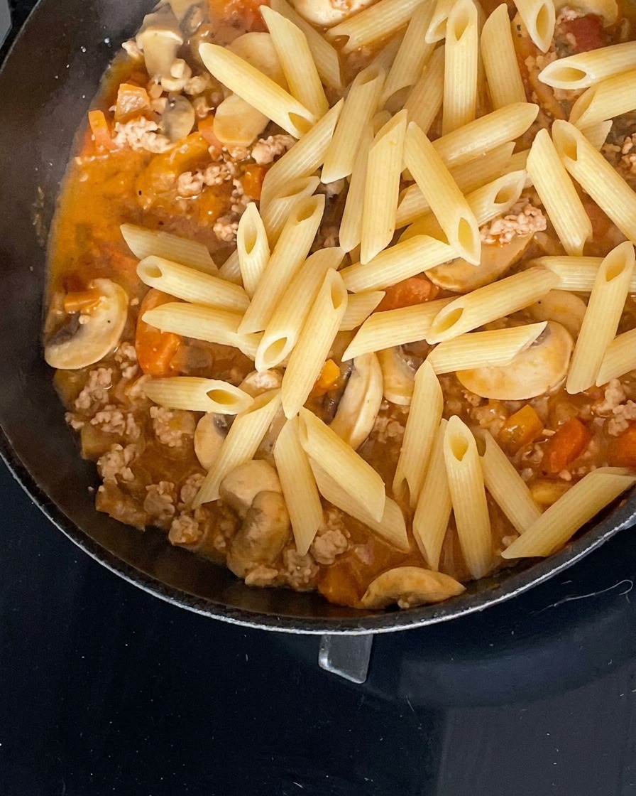 Pasta penne cocida reposando sobre la salsa en una sartén negra.