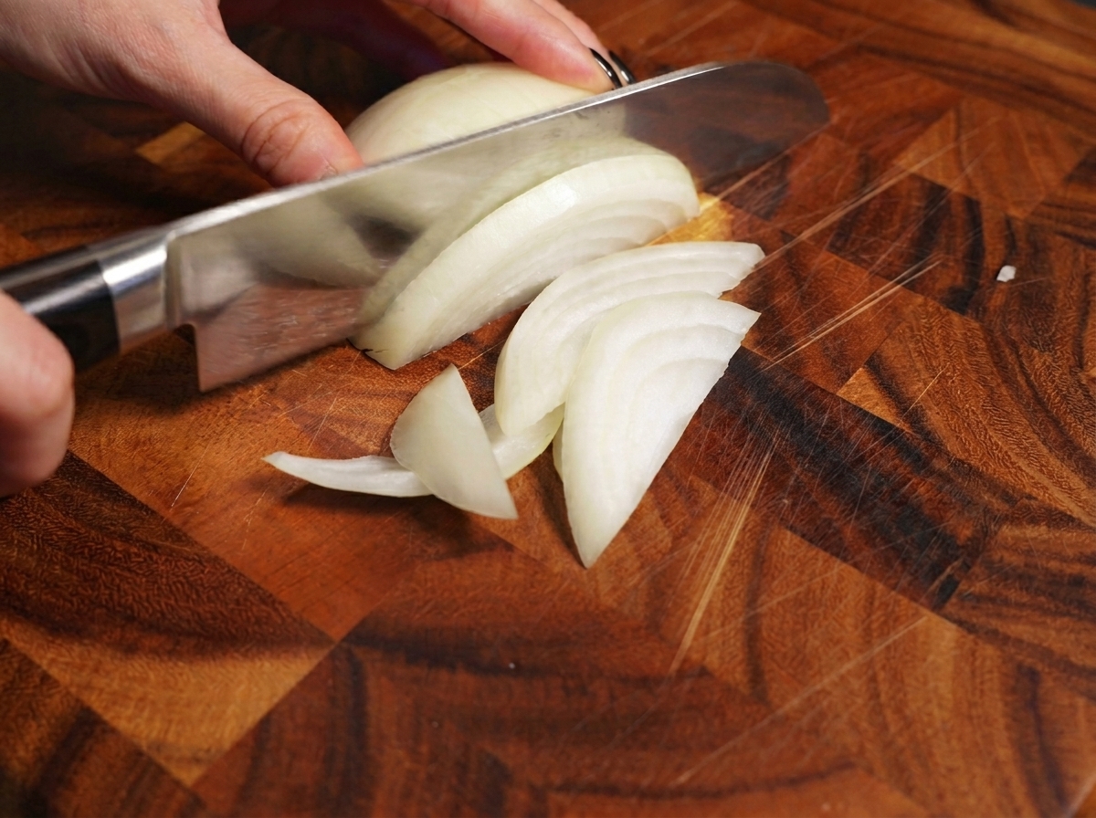 Una mano usando un cuchillo de chef para rebanar una cebolla blanca en una tabla de madera.