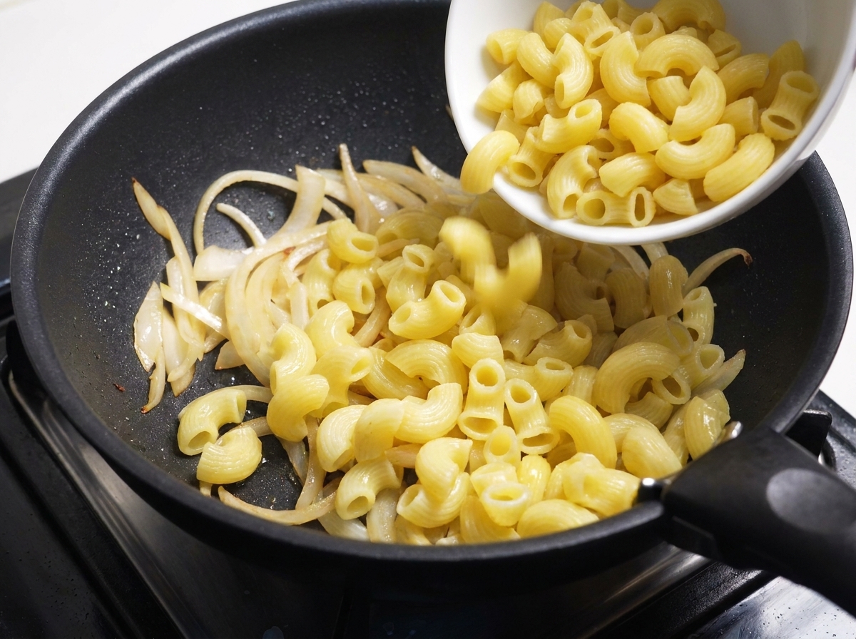 Macarrones cocidos siendo volcados desde un bol blanco a la sartén con cebollas.